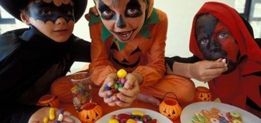 Three children in Halloween costumes sit at a table with candy and pumpkin decorations.
