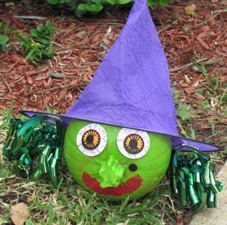 A green pumpkin decorated as a witch with a purple hat and googly eyes sits on the ground.