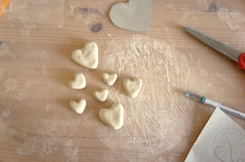 Several clay heart shapes on a wooden surface next to a craft knife and scissors.