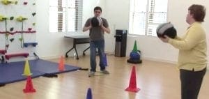 Two people are playing with medicine balls in a room with climbing walls and cones.