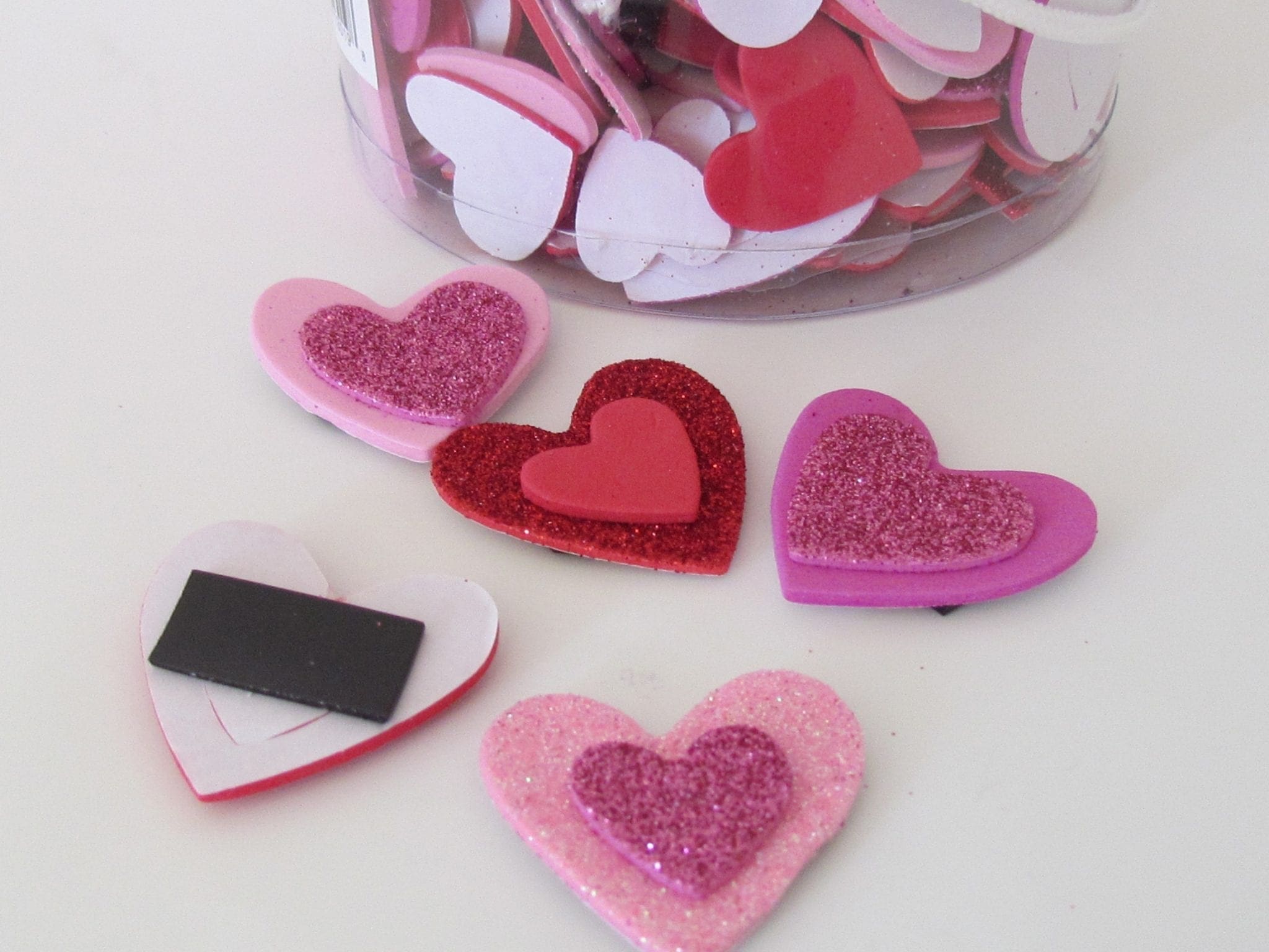 Crafted foam hearts in various shades of pink and red, some with glitter, sit on a white surface near a jar of similar heart shapes.