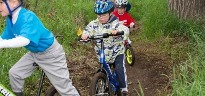 Children wearing helmets ride bicycles on a dirt path surrounded by grass and trees.
