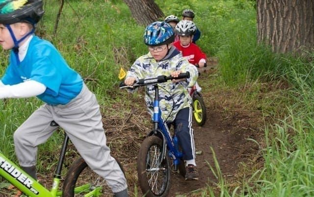 Children wearing helmets ride bicycles on a dirt path surrounded by grass and trees.