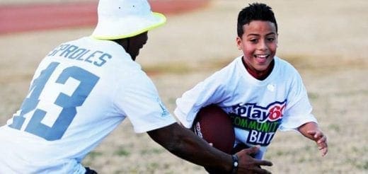 A boy smiles while holding a football as a man wearing a shirt with "PROLES 13" on it coaches him.