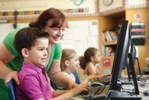 A teacher assists a young boy at a computer in a classroom.
