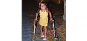 Chloe wearing a yellow dress walks with a walker.