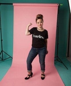 Katie Meade poses with her arm flexed in a "fearless" shirt and jeans.