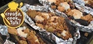 Bananas in tinfoil boats topped with melted marshmallows, chocolate, and crumbled graham crackers on a baking sheet.