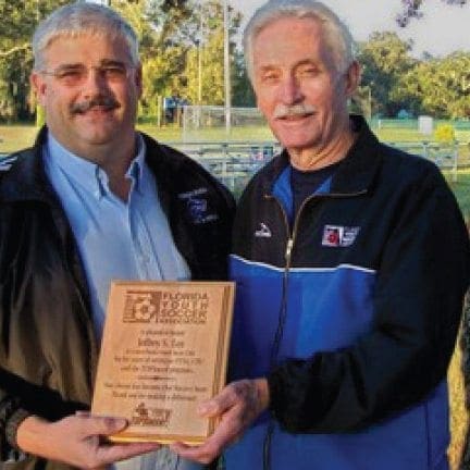 Dean Frost presents a plaque to another man at a soccer field.