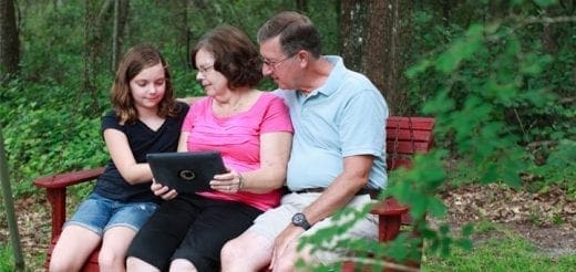 Girl and grandparents look at a tablet computer on a park bench.