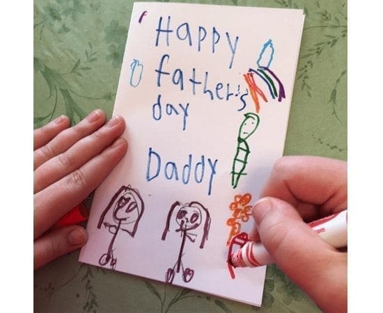 A child's hands hold a card with "Happy Father's Day Daddy" written on it, alongside stick figure drawings.