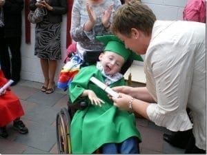 A child in a wheelchair wearing a graduation cap and gown receives a certificate.