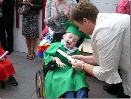 A child in a wheelchair wearing a graduation cap and gown receives a certificate.