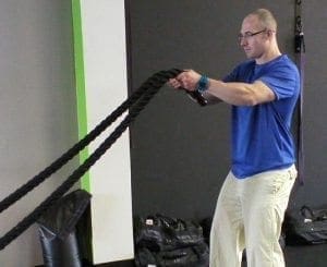 Man in blue shirt and khakis swings two thick ropes in a gym setting.