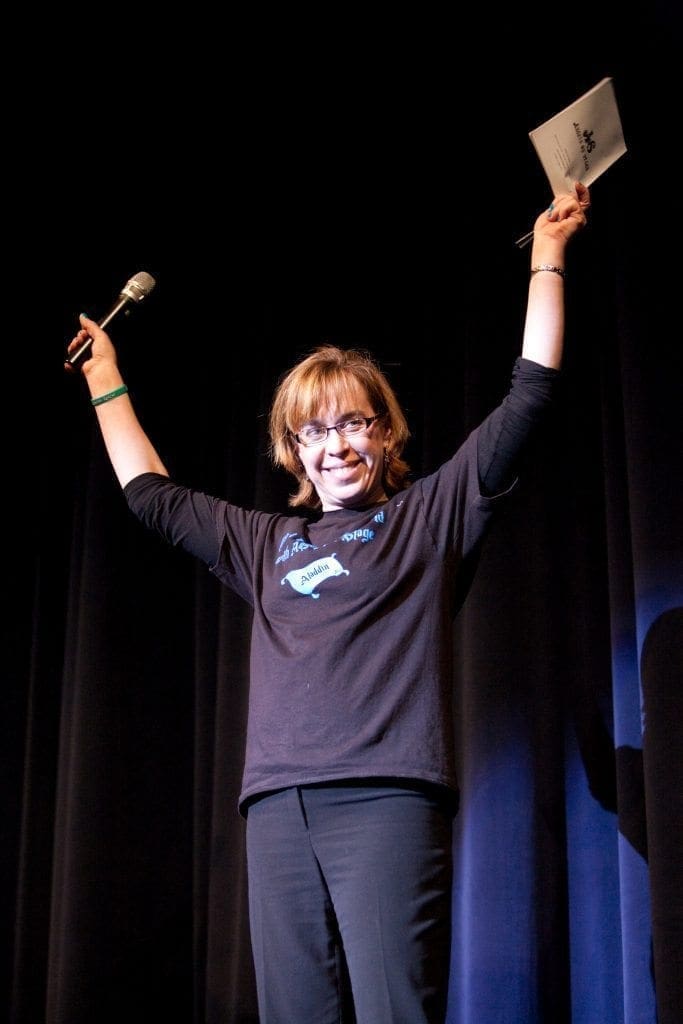 A person holds a microphone in one hand and a card in the other, with arms raised in celebration on a stage.