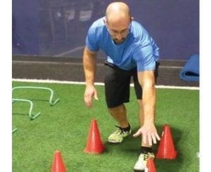 A man in athletic wear demonstrates a cone drill on an artificial turf field.