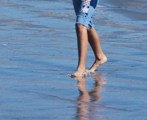 Bare feet walk through shallow water on a sandy beach.