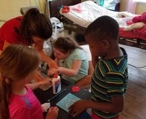 Children and adult engage in an art activity with a bottle of glue on the table.
