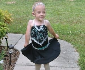 A young girl in a black ballet costume with a sparkly bodice and a green sash spins outdoors.