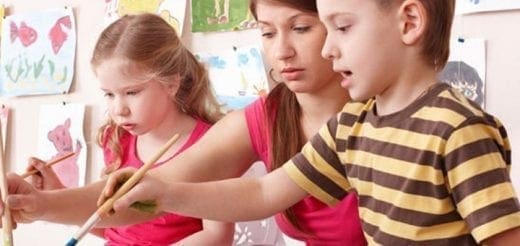 A woman helps two children paint at an easel.