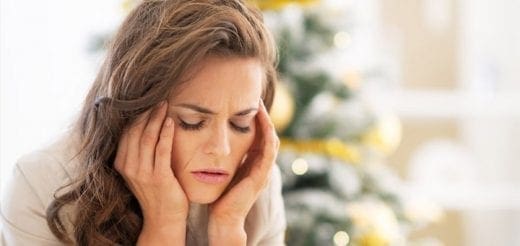 Woman with hands on temples, looking stressed in front of a decorated Christmas tree.