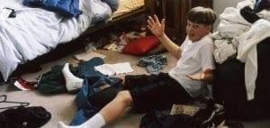 A boy sits in a cluttered room with clothes on the floor.