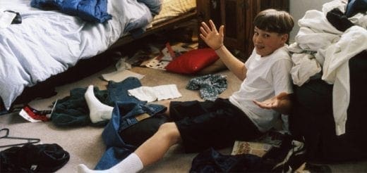 A boy sits in a cluttered room with clothes on the floor.