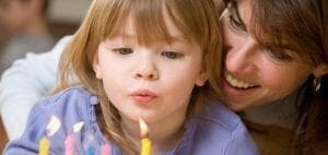 A child blows out birthday candles with a woman smiling behind.