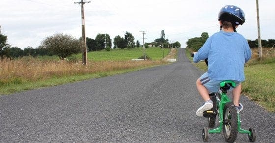 Child wearing a blue shirt and helmet rides a green bicycle with training wheels down a paved road.