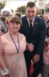 Tim Tebow in a suit with a young woman wearing a tiara and a pink dress.