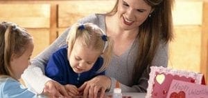 A woman smiles as she helps two young children make Valentine's Day cards.