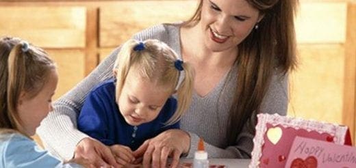 A woman smiles as she helps two young children make Valentine's Day cards.