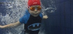Child in red swim cap and goggles underwater in a pool.