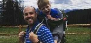 A father smiles while carrying his son in a backpack carrier through a wooded area.