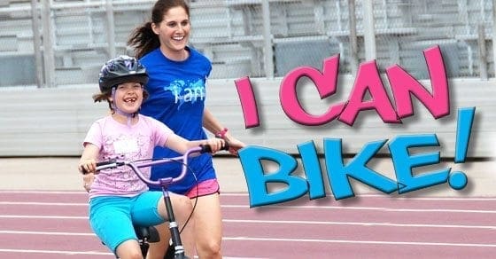A young girl wearing a helmet smiles while riding a bike as a woman runs beside her.