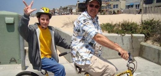 Two people wearing helmets ride bicycles near a beach.