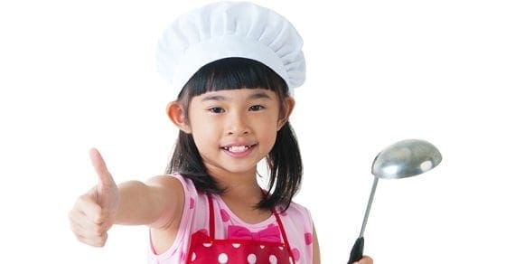 Child in chef hat and apron gives a thumbs up while holding a ladle.