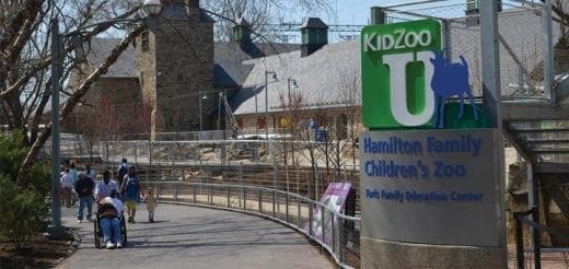Entrance sign for KidZooU Hamilton Family Children's Zoo & Faris Family Education Center with visitors on a pathway.