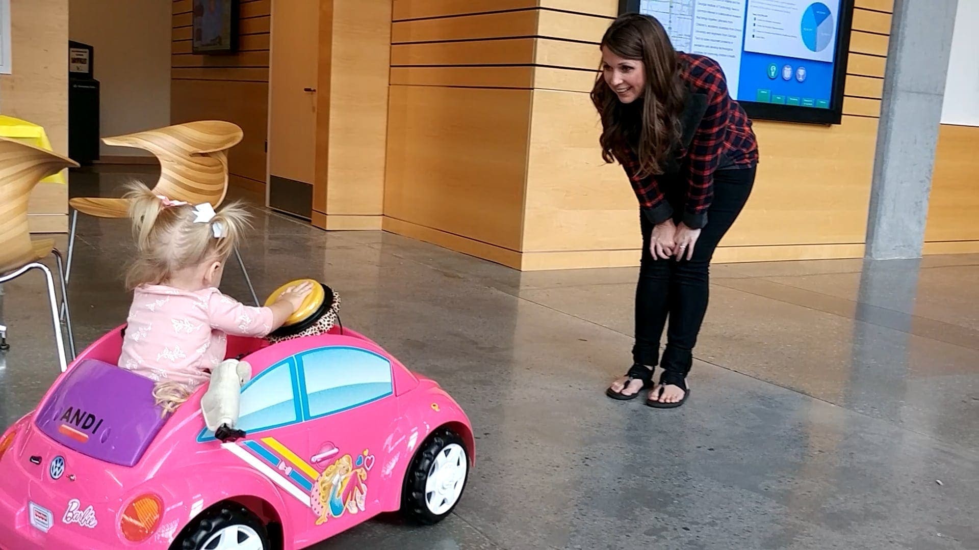 A woman kneels and smiles at a toddler in a pink toy car.