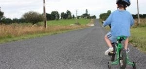 Child riding a green bicycle with training wheels on a gravel road.