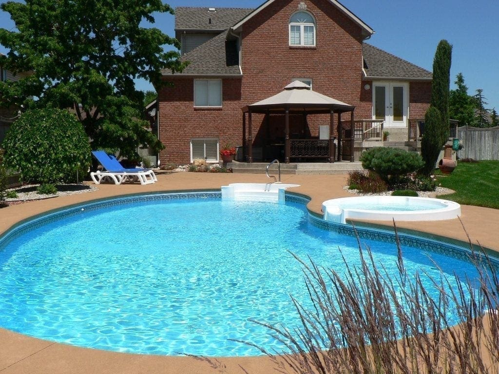An outdoor swimming pool and hot tub in front of a large brick house with a gazebo.