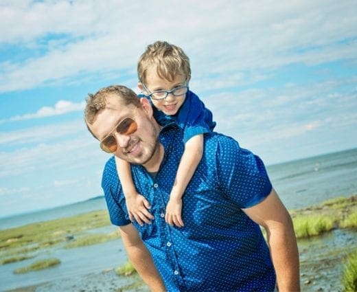 A father wearing sunglasses gives his son a piggyback ride outdoors.