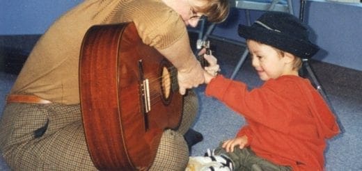 A child in an orange shirt and black hat holds hands with an adult holding a guitar.