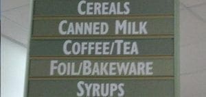 Sign with rows listing "Cereals," "Canned Milk," "Coffee/Tea," "Foil/Bakeware," and "Syrups.