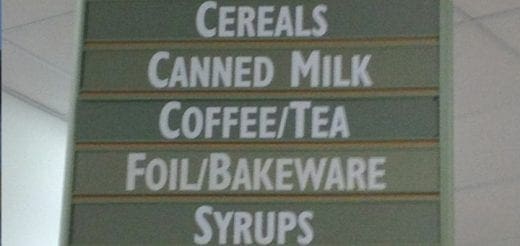 Sign with rows listing "Cereals," "Canned Milk," "Coffee/Tea," "Foil/Bakeware," and "Syrups.