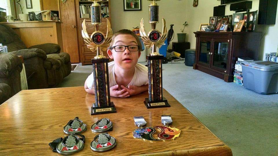 A person with glasses looks at the camera between two trophies, with medals on the table in front.