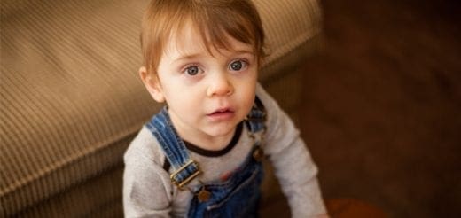 Toddler wearing denim overalls sits near a couch.