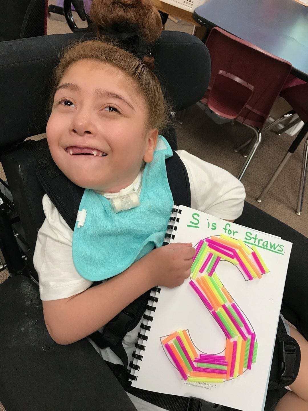 A young person smiles while holding a notebook with the letter S made of colorful straws.