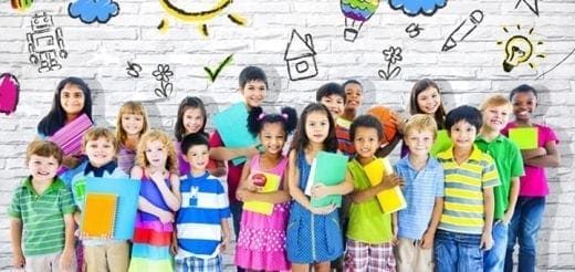 A diverse group of children stand in front of a brick wall with chalk drawings of school supplies and the sun.