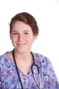 A young woman in scrubs and a stethoscope smiles.
